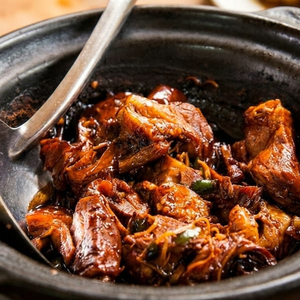 Braised meat in a dark, rustic clay pot with a serving spoon, showcasing rich brown sauce.